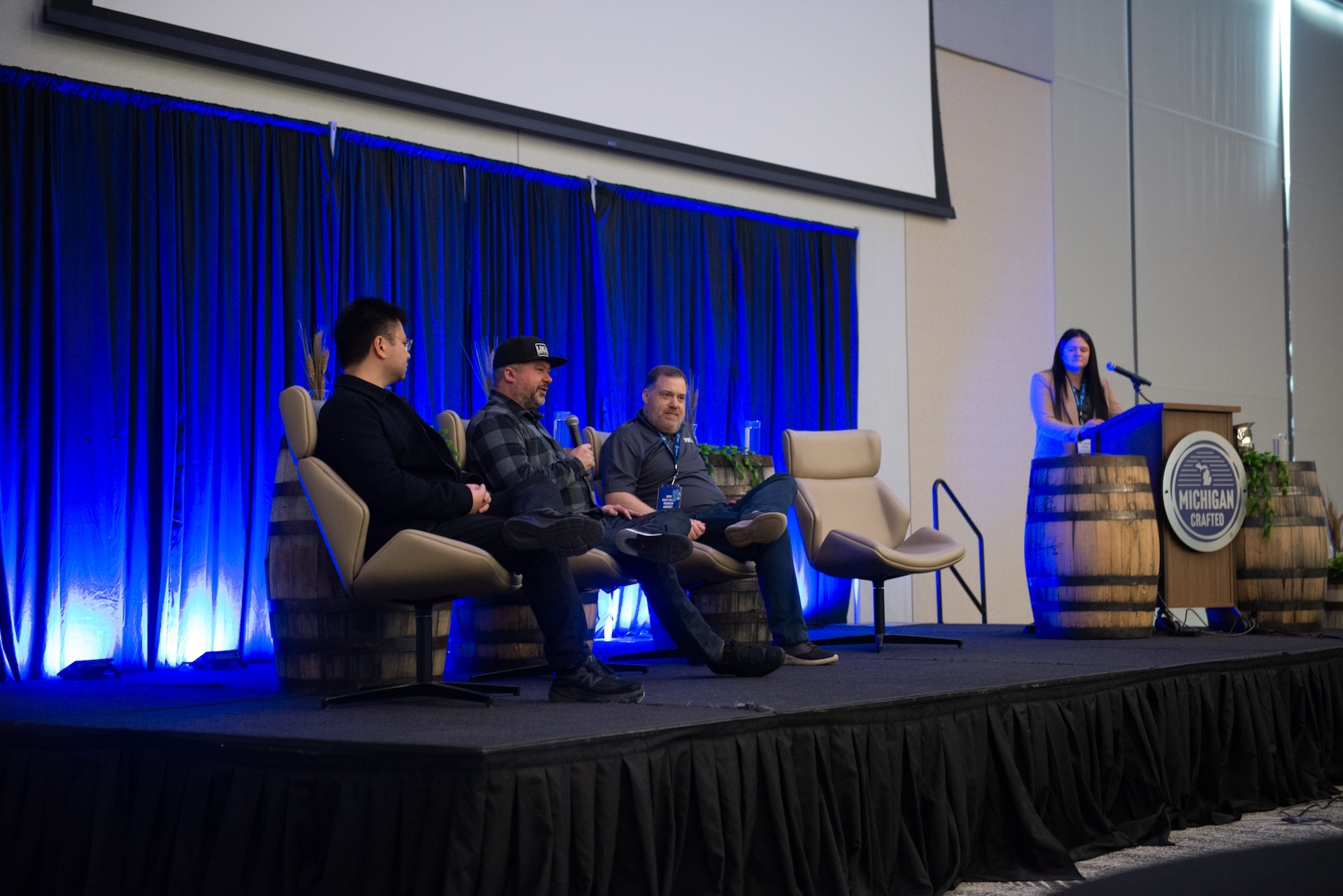 Conference panel with three people seated on a stage, and one person speaking behind a podium with 'MICHIGAN CRAFTED' signage